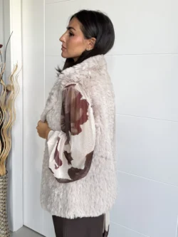 Mujer con chaleco beige de pelo sintético visto en ángulo tres cuartos, combinando con blusa de gasa estampada.